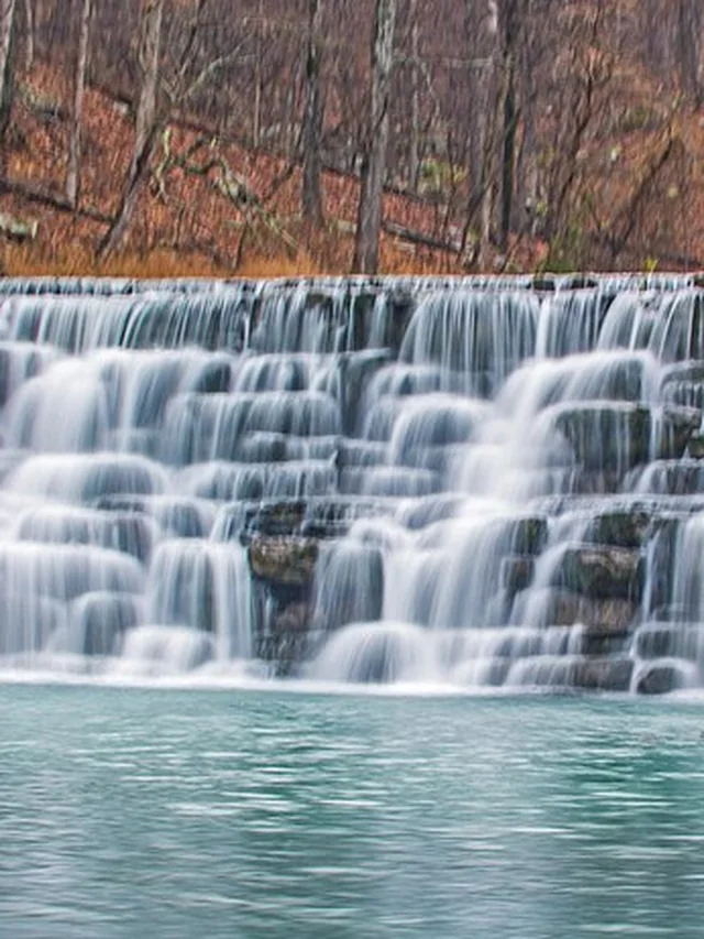 A wide waterfall spilling over layered rock into a green pool at Devil's Den State Park